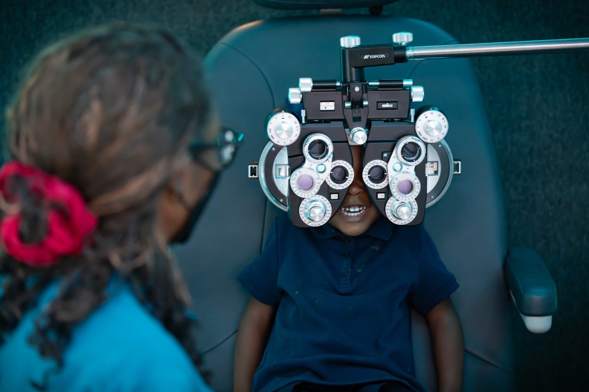A child in a dark blue polo shirt sits in an optometry chair and looks through a phoropter.