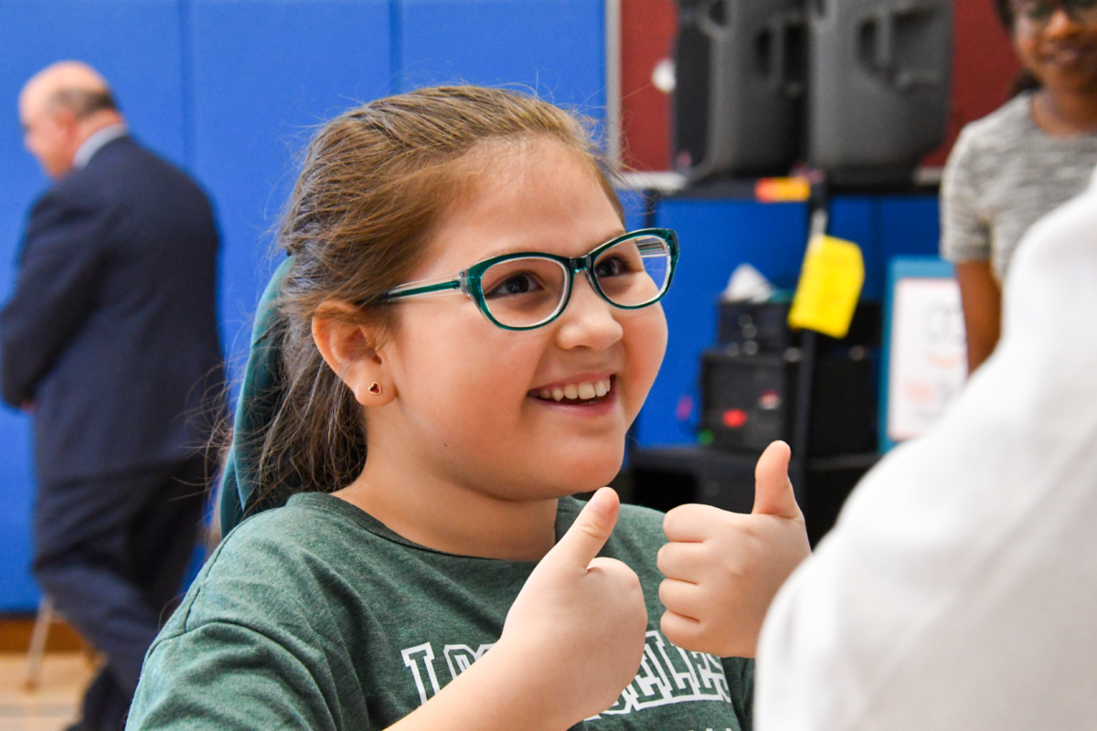 A 5th grade student wearing a green shirt and green framed glasses gives a double thumbs up.