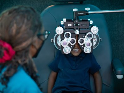 A child in a dark blue polo shirt sits in an optometry chair and looks through a phoropter.