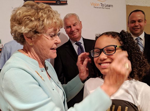 Sally Gore, Chair of Vision To Learn’s Delaware Advisory Board, puts glasses on a child as Wilmington Mayor Michael Purzycki and others look on.