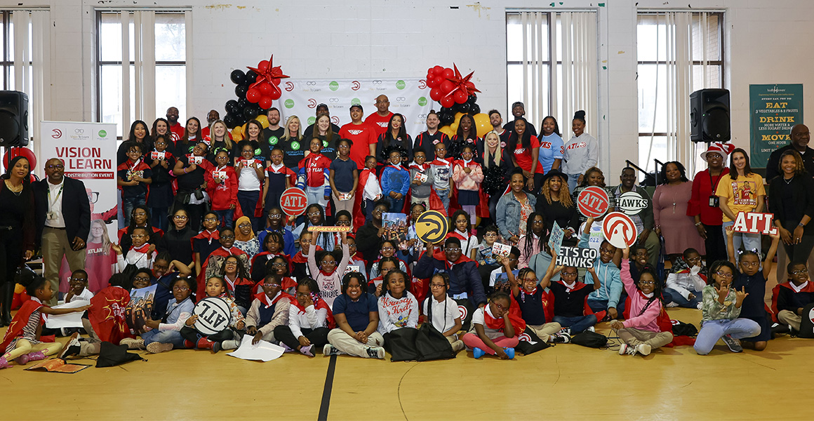 A large group of young students, and representatives from Vision To Learn and the Atlanta Hawks, posing for group picture against a white wall in a school gymnasium.