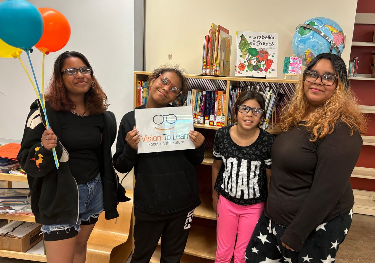 Four young women wearing glasses pose for a picture. The woman on the far left is holding three balloons and the woman to her right is holding a sign with Vision To Learn's logo on it.