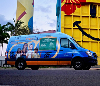 A blue Vision To Learn clinic with pictures of four children on the side sits in a parking lot in front of a building whose yellow wall has a smiley face painted on it.