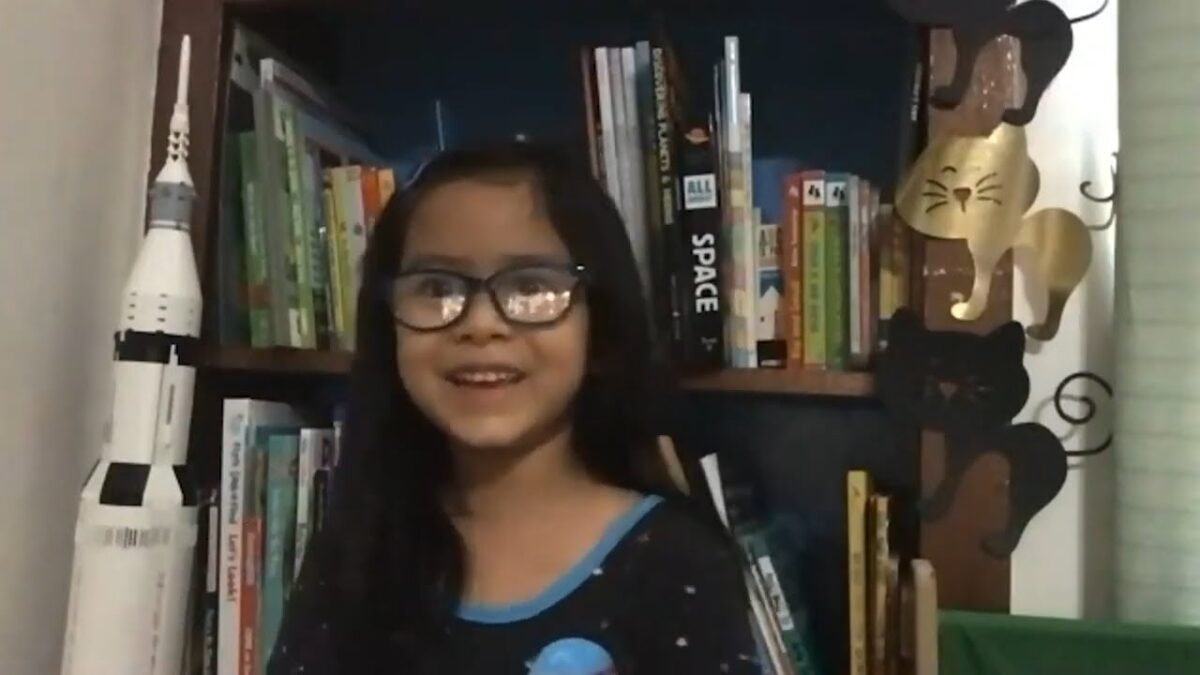 A young girl wearing glasses stands in front of a bookshelf with a model of Apollo 11 leaning against it.