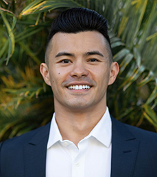 A man wearing a blue suit jacket with a white dress shirt stands in front of foliage.