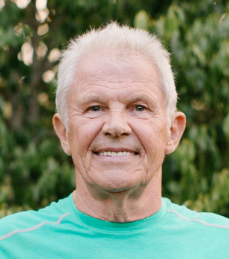 A man in a teal crew neck shirt stands in front of foliage. 