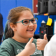 A 5th grade student wearing a green shirt and green framed glasses gives a double thumbs up.