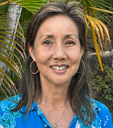 A woman in a floral blue button up shirt stands in front of foliage.
