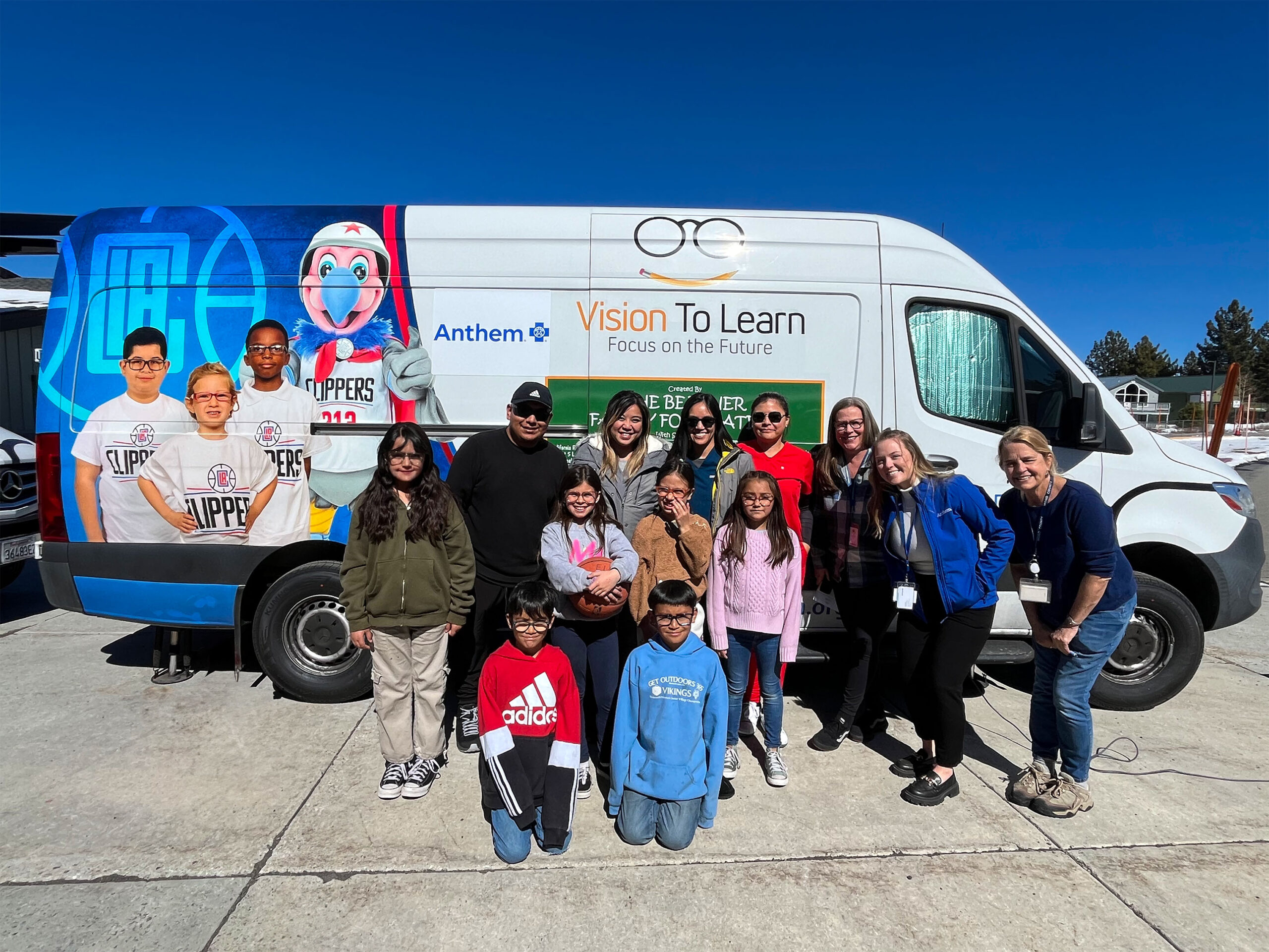 A group of six students pose for a picture with seven representatives from Anthem Blue Cross while in front of a Vision To Learn clinic with LA Clippers branding.