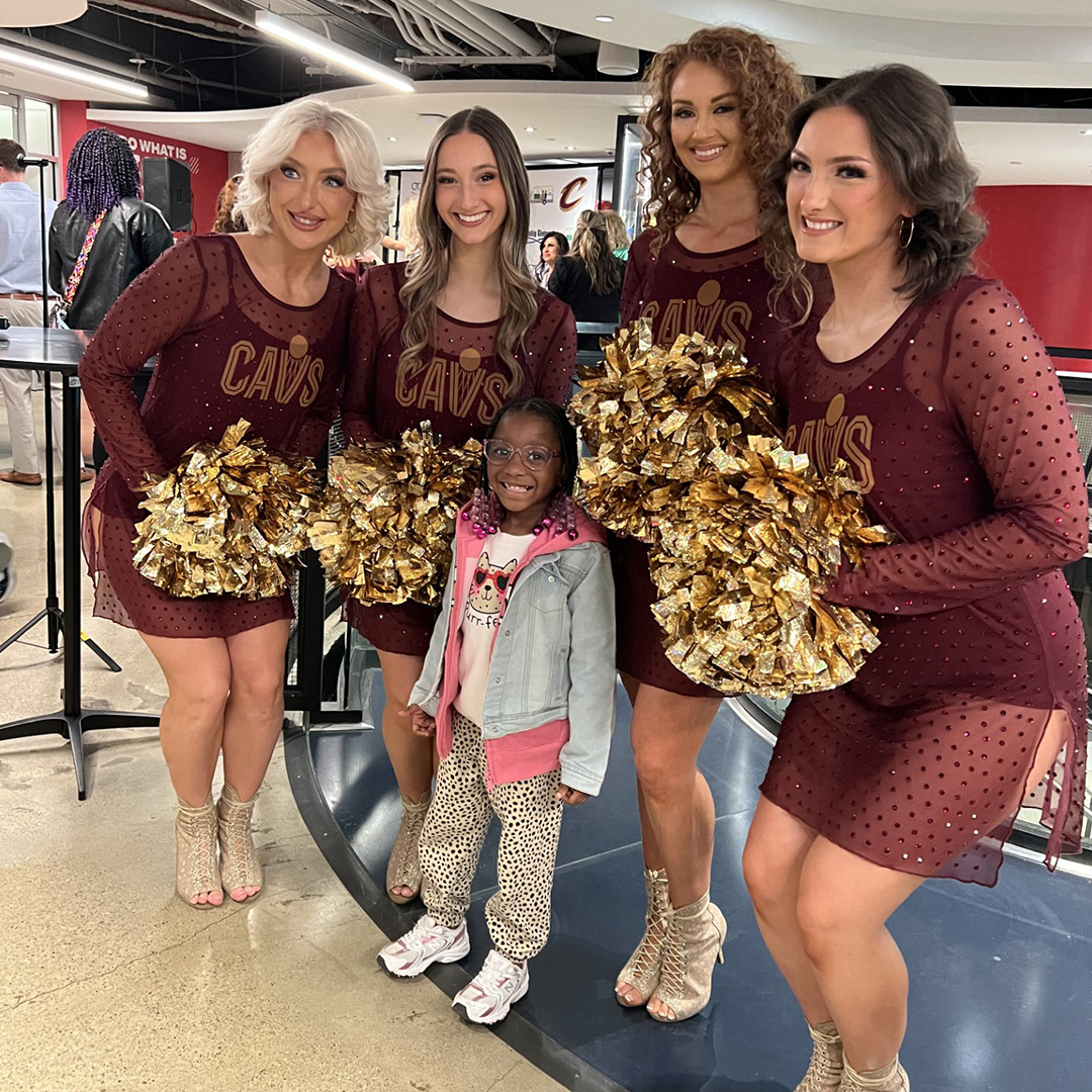 A young girl in a denim jacket is flanked by four cheerleaders in sheer burgundy dresses carrying golden pom poms.