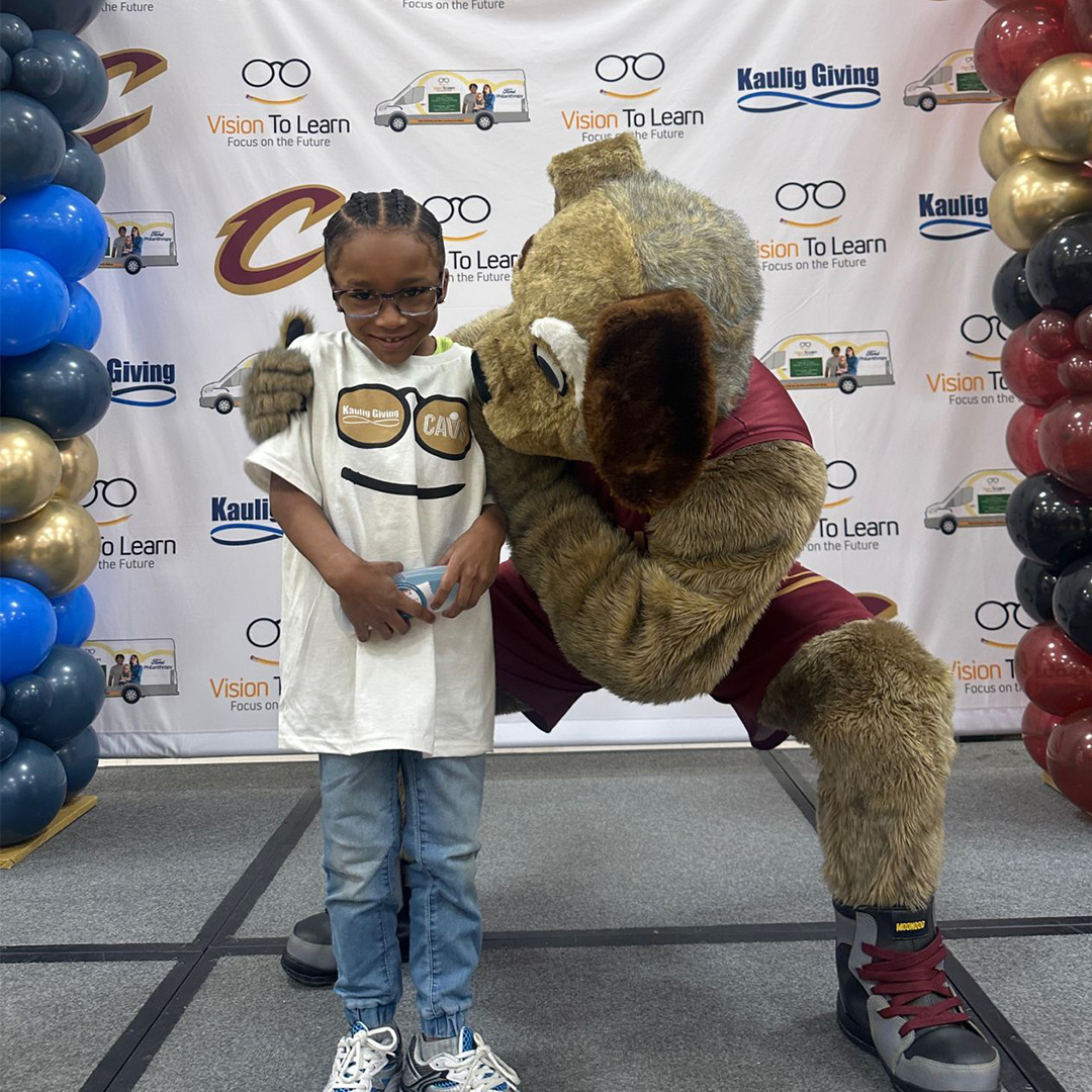An elementary school student in a white t-shirt poses for a picture with a large dog mascot wearing a red jersey.