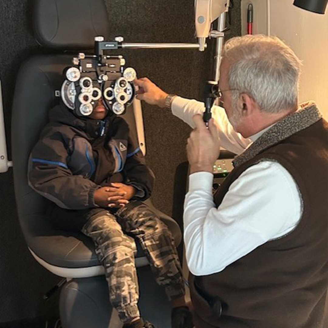 A boy in camouflage pants and a black winter coat sits in an optometry chair with a phoropter in front of his face as an optometrist conducts an eye exam.
