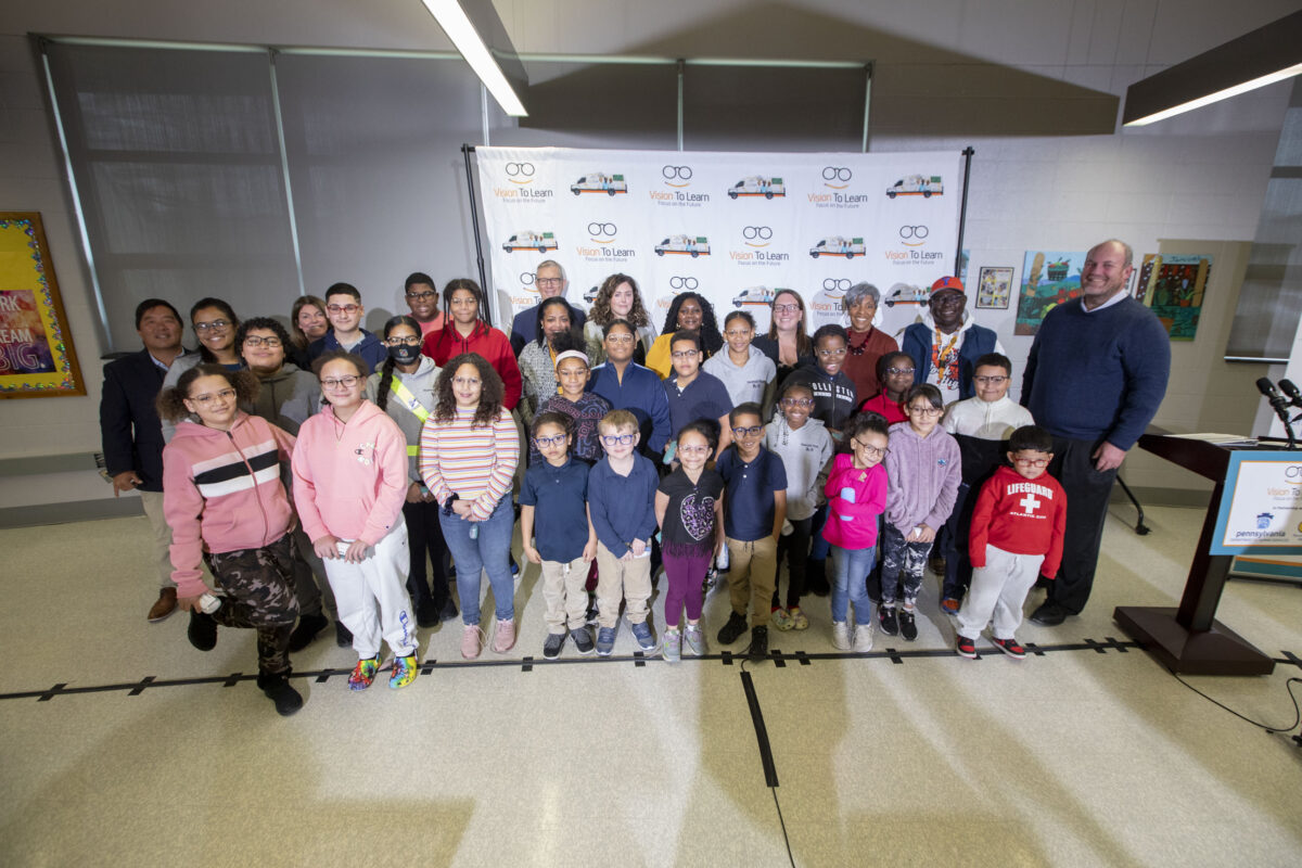 Elementary school students pose for a group picture with their new glasses while standing in front of a Vision To Learn backdrop.