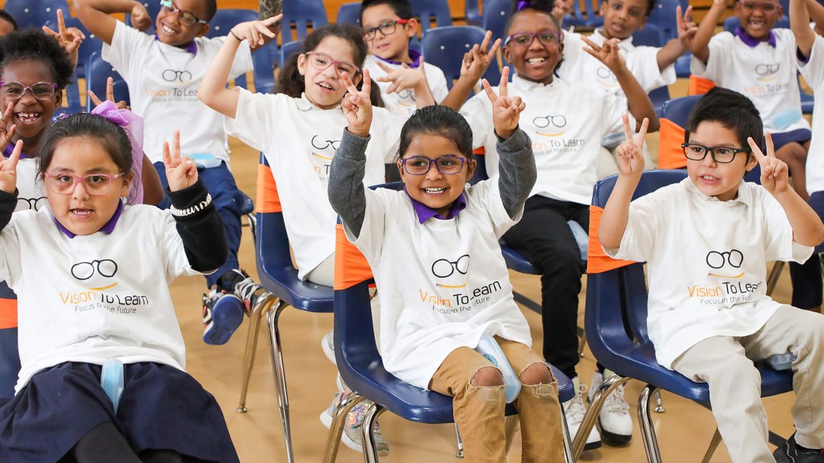 A group of students wearing white t-shirts featuring Vision To Learn's logo, sit on blue chairs in the middle of a school' gymnasium.