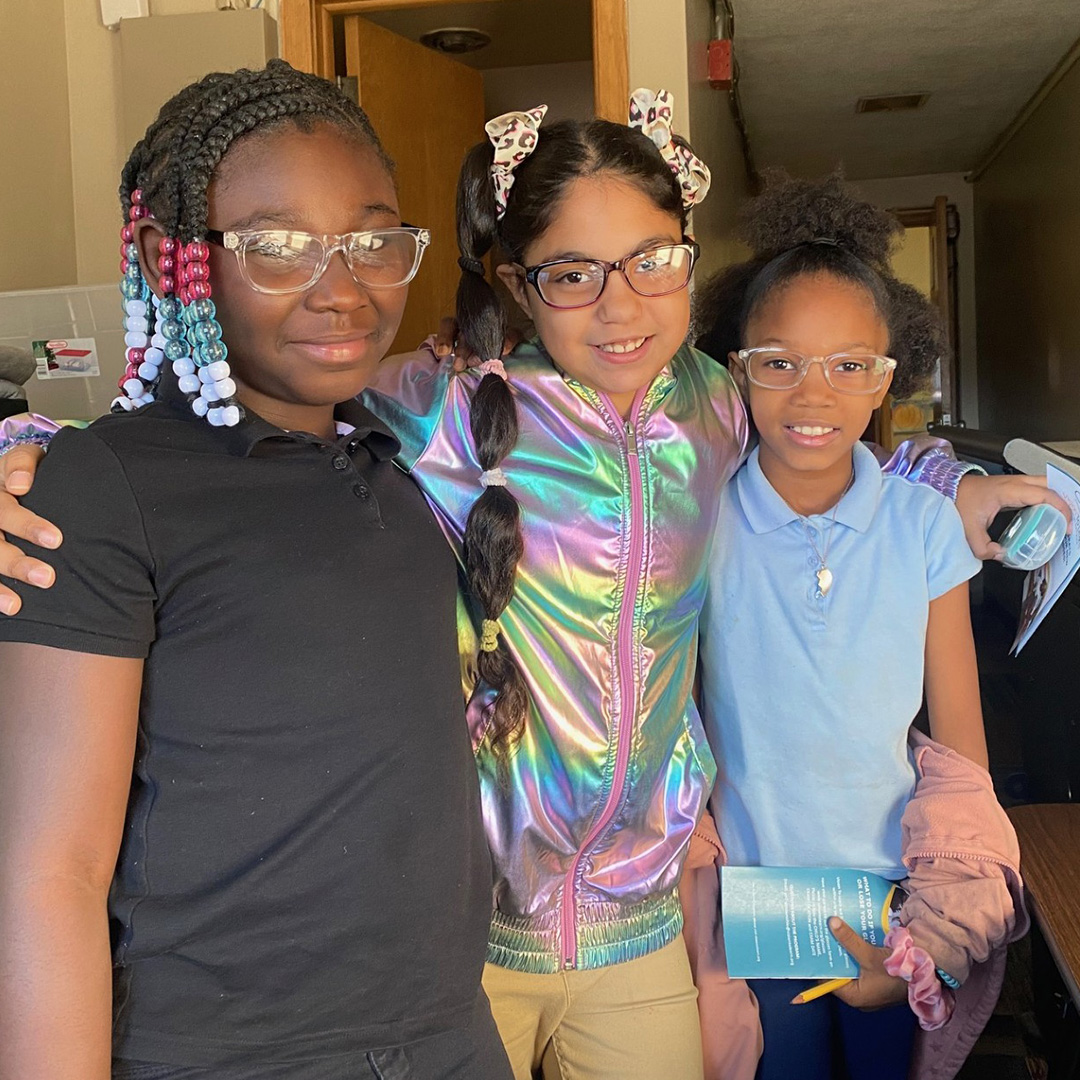 Three young girls pose for a picture together in front of the entrance to a classroom.