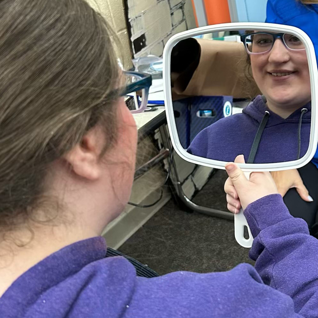 A young woman in a purple hoodie looks at her glasses in a handheld mirror.