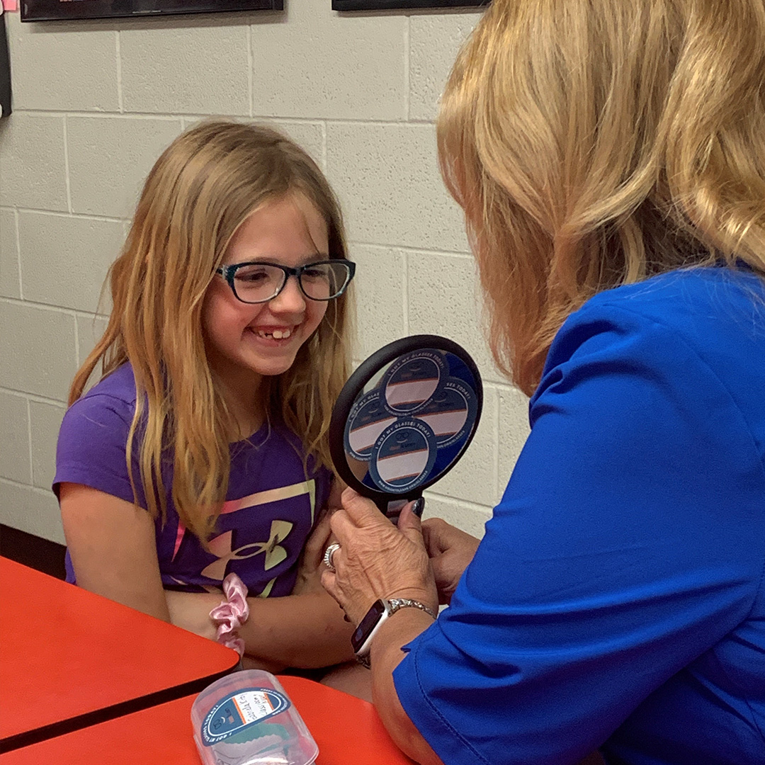 A young girl sits at a red table in front of an optician in blue scrubs who is holding a handheld mirror for her to look at.