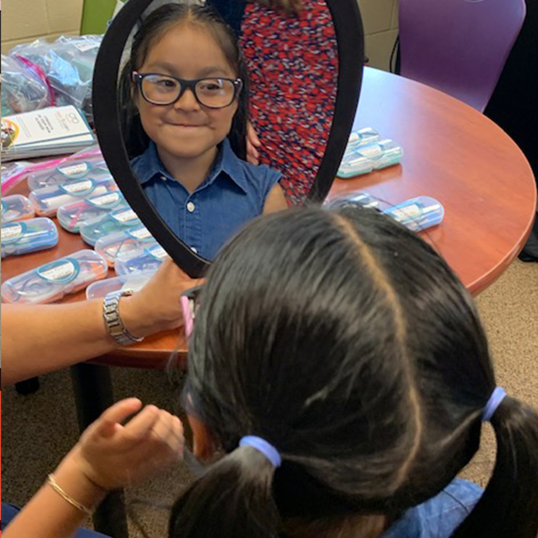 A young girl wearing a blue denim collared shirt looks in a mirror at her glasses.