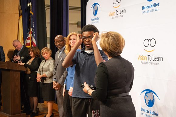 Franklin Walker, Chief Schools Administrator of Jersey City Public Schools, and Laura Overdeck, Chair of the Overdeck Family Foundation, look on while a Frank R. Conwell student receives his glasses.