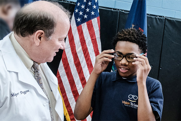 Detroit Mayor Mike Duggan helps an elementary school student in a Vision To Learn polo shirt try on a pair of glasses.